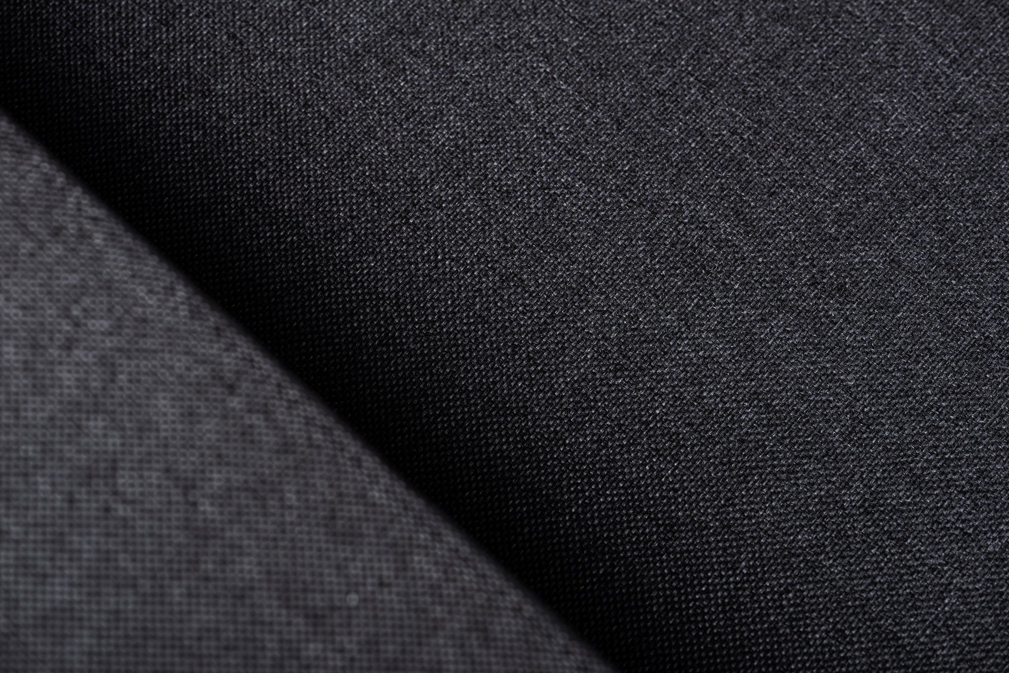 Close-up van donkergrijze stof met een fijn, gestructureerd weefsel. Het oppervlak vertoont een subtiele glans, wat de gladheid en details van het materiaal benadrukt. Diagonale schaduwen creëren diepte en contrast over de vouwen van de stof.