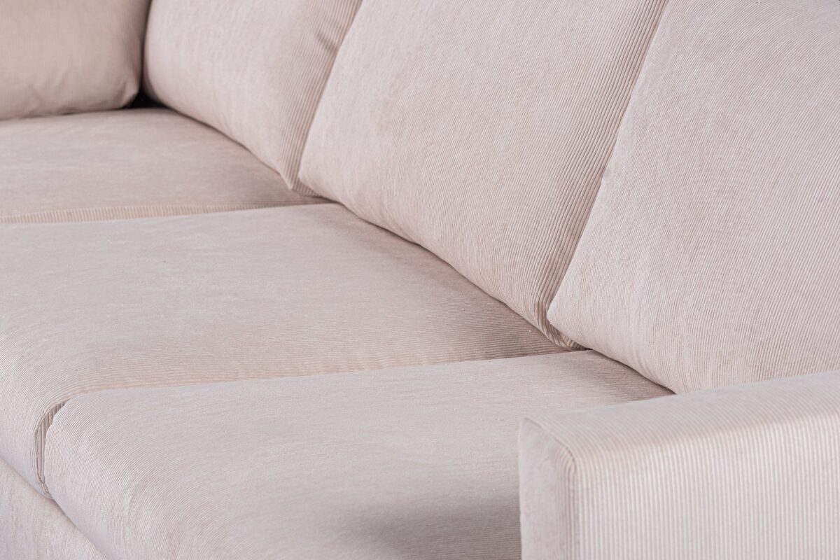 Close-up van een beige, met stof beklede bank met drie gewatteerde zittingen en een licht zichtbare armleuning. De stof heeft een subtiele textuur en de kussens lijken zacht en comfortabel.