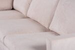 Close-up van een beige, met stof beklede bank met drie gewatteerde zittingen en een licht zichtbare armleuning. De stof heeft een subtiele textuur en de kussens lijken zacht en comfortabel.