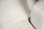 Close-up van een beige stoffen bank met textuur, met een hoek waar twee kussens elkaar ontmoeten. De stof heeft een subtiel weefpatroon, waardoor het oppervlak een licht ruw, tastbaar uiterlijk krijgt. De neutrale kleur creëert een zachte, uitnodigende look.