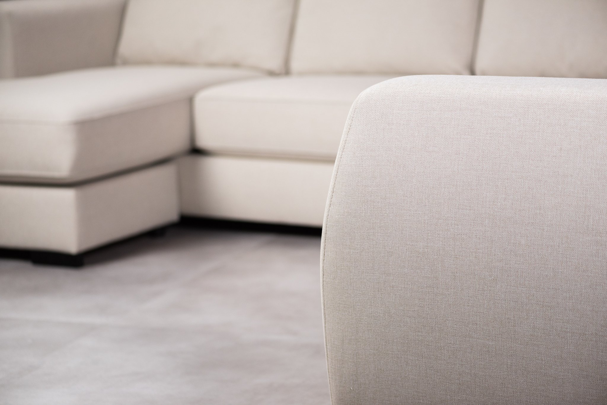 Close-up van een beige sectionele bank met wazige achtergrond. De textuur van de stof is zichtbaar, waardoor de bekleding wordt benadrukt. De zitopstelling bestaat uit een chaise longue en meerdere kussens, geplaatst op een lichtgekleurde vloerbedekking.