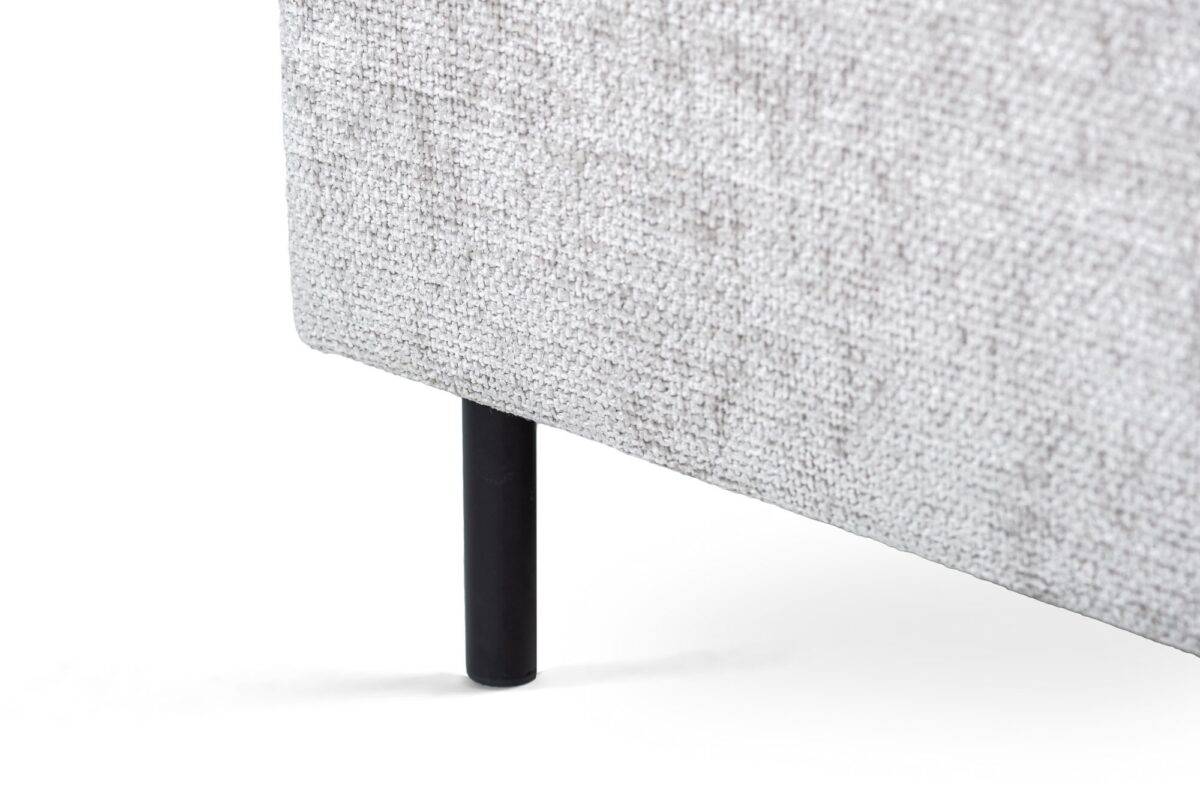 Close-up van een lichtgrijs gestoffeerd meubelstuk met een zwarte cilindrische poot linksonder. De stof heeft een getextureerde, geweven uitstraling. De achtergrond is effen wit.