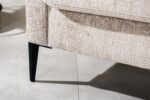 Close-up van een beige getextureerde hoekbank met een zwarte taps toelopende poot, geplaatst op een lichtgekleurde betegelde vloer. De stof heeft een geweven uiterlijk en de verlichting werpt een schaduw op de vloer.