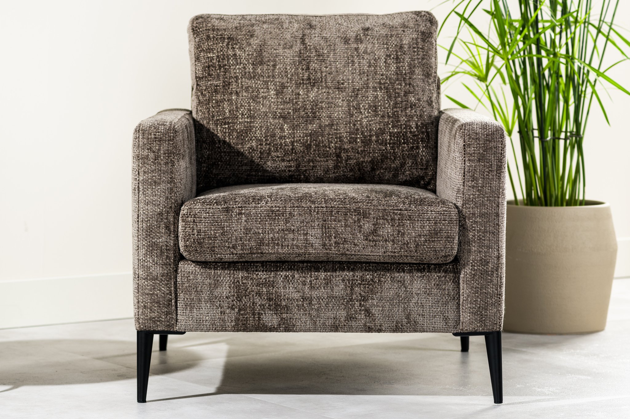 Een pluche, getextureerde bruine fauteuil met zwarte poten staat op een lichtgekleurde vloer. Een grote, groene potplant staat rechts, tegen een gebroken witte muur, wat een gezellige en uitnodigende sfeer creëert.