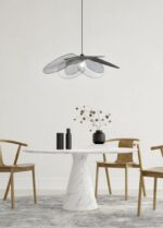 Een minimalistische eethoek met een ronde witte marmeren tafel, twee houten stoelen en een zwarte vaas met gedroogde bloemen. Een moderne zwarte plafondventilator met een gebogen design hangt erboven. De setting is tegen een effen witte muur.