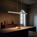 Een moderne eetkamer met een houten tafel en een fruitmand in het midden. Een strakke, langwerpige lamp hangt erboven. De kamer heeft zachte, warme verlichting, een houten dressoir met decoratieve items en een groot raam op de achtergrond.