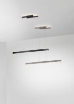 Twee moderne, minimalistische hanglampen hangen aan het plafond. De ene is zwart en de andere lichtgrijs. Beide hebben slanke, rechthoekige vormen en hangen aan dunne kabels, waardoor er zacht licht op het witte plafond valt.