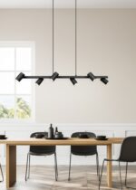 Een moderne eetkamer met een houten tafel voor zes personen, met zwarte stoelen en servies. Boven ons hoofd hangt een strakke zwarte kroonluchter met zes spots. Een groot raam biedt uitzicht op de bomen buiten en de kamer heeft een lichte, luchtige sfeer.