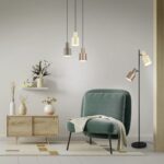 Een moderne woonkamer met een grote groene fauteuil, een houten kast en een hoge staande lamp. Hanglampen hangen erboven. Decoratieve items en planten staan op de kast en wandplanken, met een lichtgekleurd geometrisch kleed op de vloer.