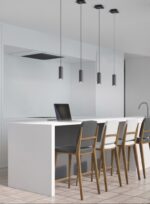Minimalistische keuken met een strak wit eiland, met vier houten krukken met grijze en witte kussens. Vier hanglampen hangen boven het eiland en een laptop staat open op het aanrecht. De vloer is betegeld in een licht patroon.