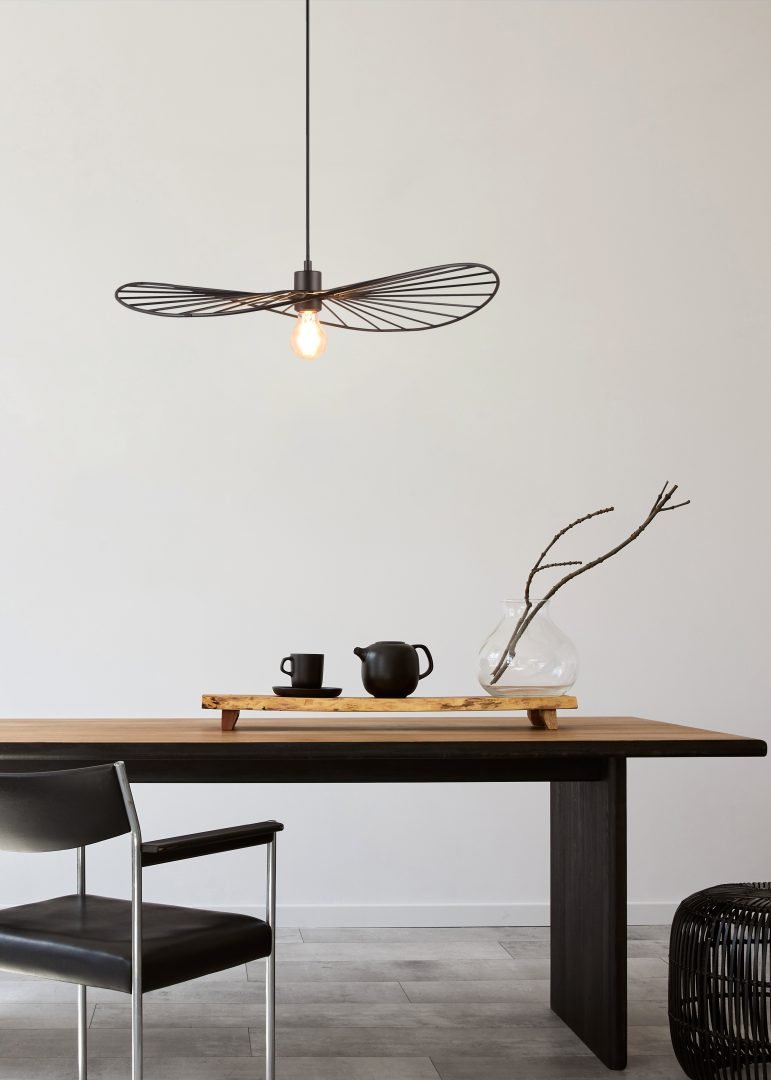 Minimalistische eethoek met een houten tafel, een enkele zwarte stoel en een unieke hanglamp met draadframe erboven. De tafel heeft een theepot op een houten dienblad en een glazen vaas met takken. Lichtgrijze muren vormen de achtergrond.