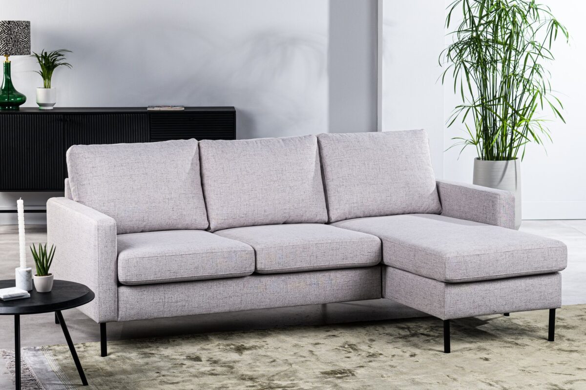 Een moderne woonkamer met de 3 zits bank CL L+R in de stof Valente V311 grijs, gecombineerd met een zwarte ronde salontafel met daarop kleine plantjes en een kaars, een beige vloerkleed, zwart dressoir, hoge groene plant en witte muren.
