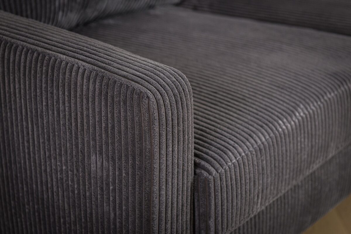 Close-up van een donkergrijze, geribbelde stoffen fauteuil, met het textuurpatroon op de armleuning en zijkant. De bekleding geeft een gevoel van zachtheid en comfort.