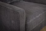 Close-up van een donkergrijze, geribbelde stoffen fauteuil, met het textuurpatroon op de armleuning en zijkant. De bekleding geeft een gevoel van zachtheid en comfort.