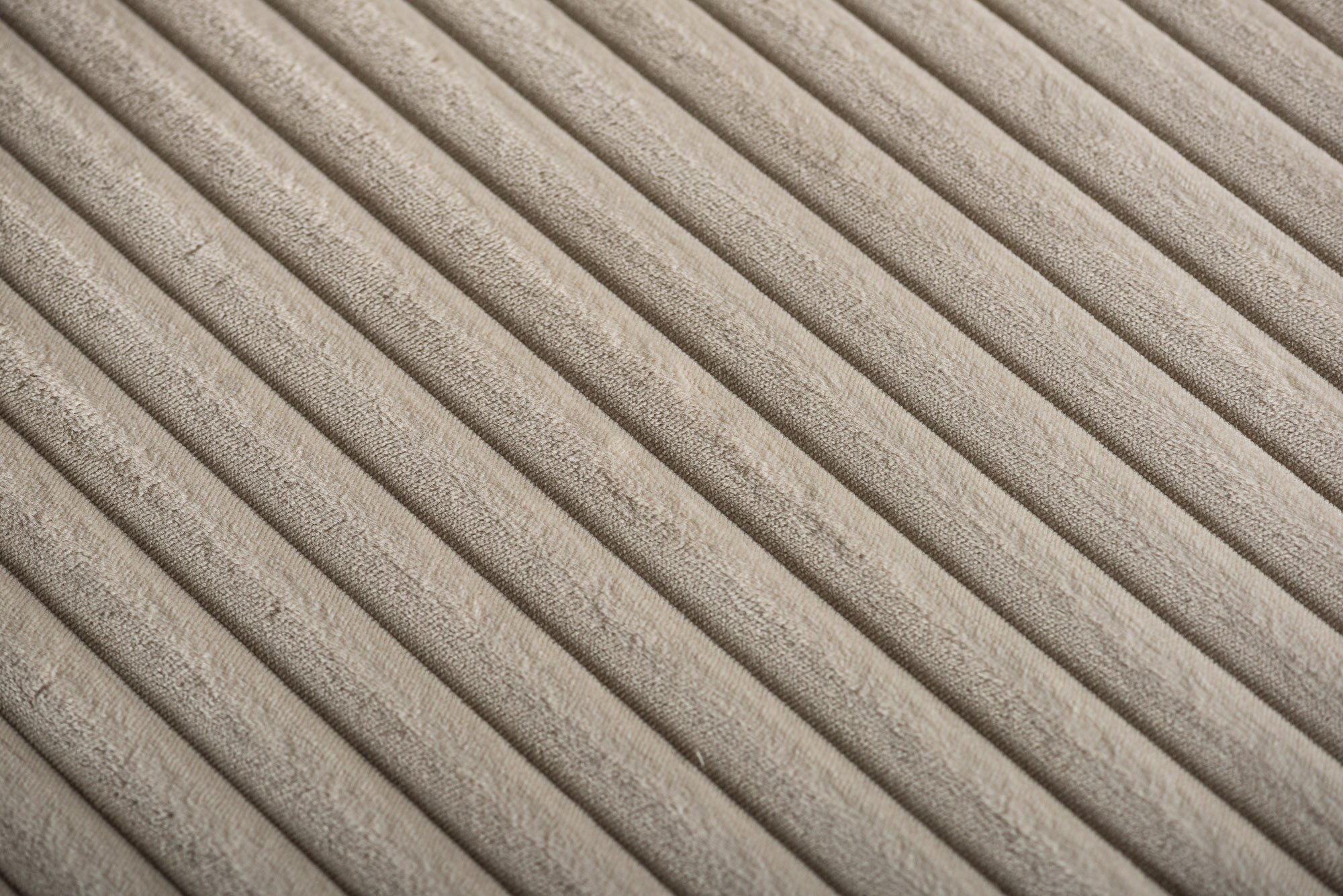 Close-up van een beige geribbelde stof met gelijkmatig verdeelde verticale ribbels. De textuur lijkt zacht en licht pluizig, wat duidt op natuurlijke vezels. Het perspectief creëert een gevoel van diepte en dimensie.