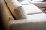 Close-up van een beige stoffen bank met eenvoudige, strakke lijnen en rechthoekige kussens. De bank baadt in zacht natuurlijk licht, waardoor de textuur wordt benadrukt. Een vleugje groene plant is zichtbaar in de rechterbovenhoek.