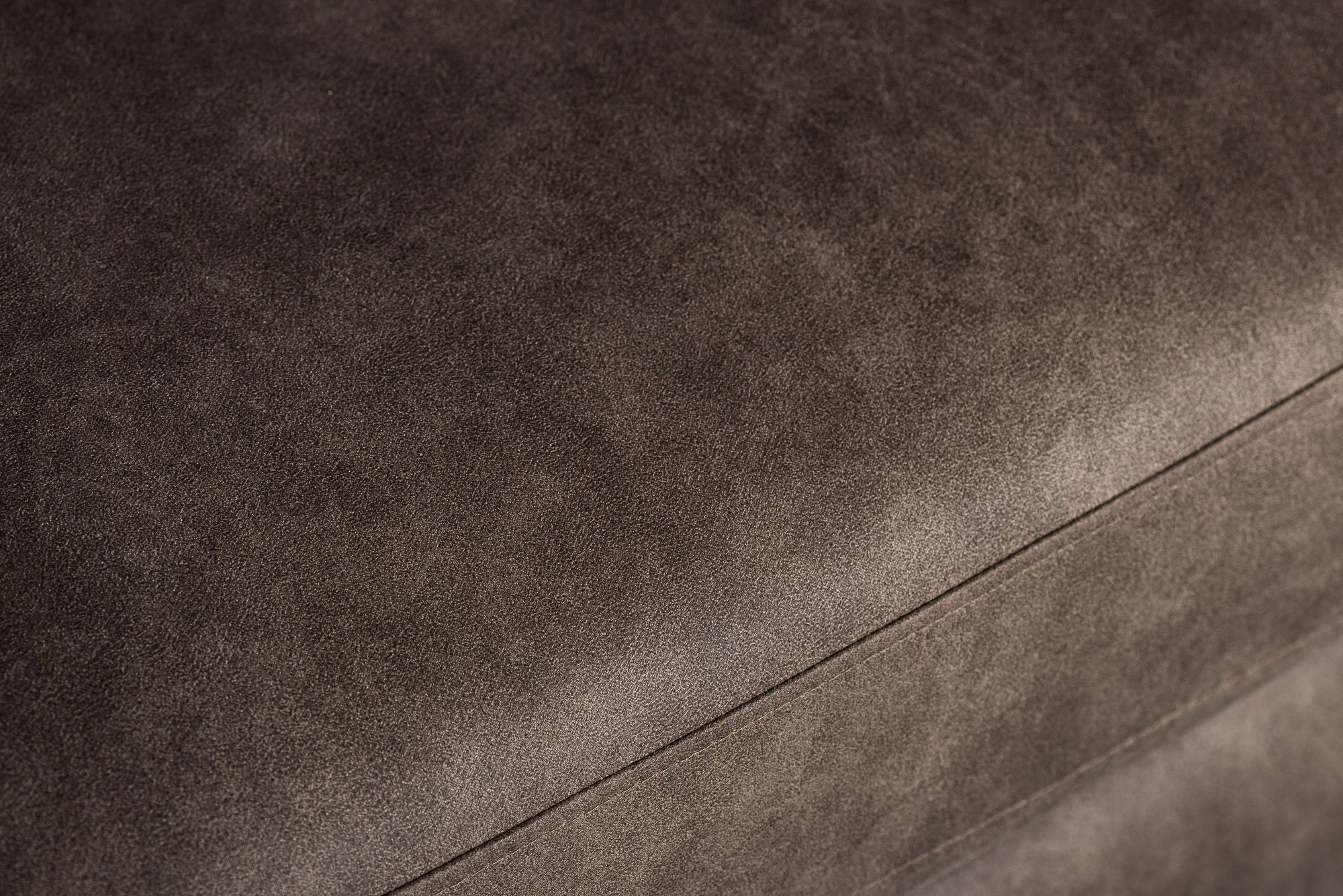 Close-up van een glad, bruin leren oppervlak met zichtbare textuur en subtiele schaduw. Het materiaal lijkt zacht en mat, met een naad die horizontaal over de afbeelding loopt.