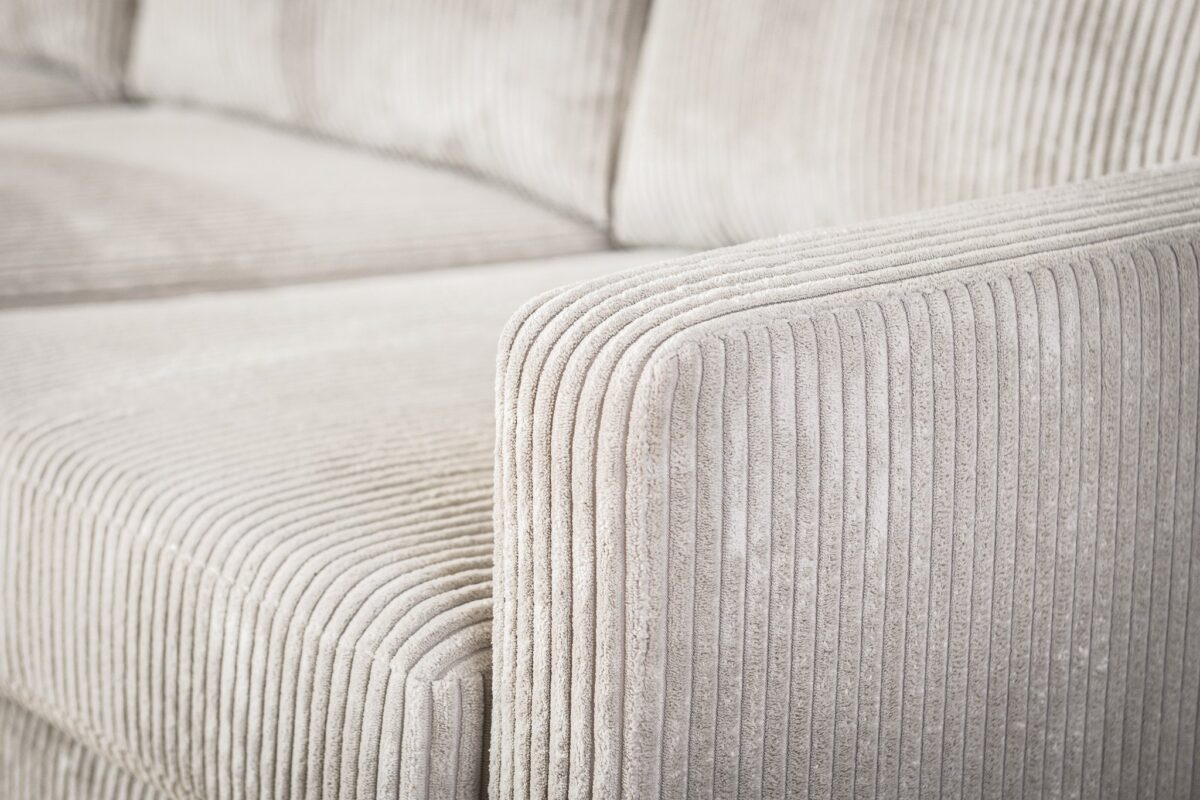 Close-up van een beige corduroy bank, met de getextureerde ribbels op de armleuning en het kussen. De stof lijkt zacht en pluche, met een subtiele glans in het licht, wat de details en patronen van het materiaal benadrukt.