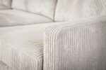 Close-up van een beige corduroy bank, met de getextureerde ribbels op de armleuning en het kussen. De stof lijkt zacht en pluche, met een subtiele glans in het licht, wat de details en patronen van het materiaal benadrukt.