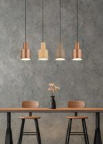 Vier hanglampen in verschillende tinten hangen boven een moderne tafel met twee houten stoelen. Een zwarte vaas met roze bloemen staat in het midden van de tafel. De achtergrond is een grijze muur met textuur.