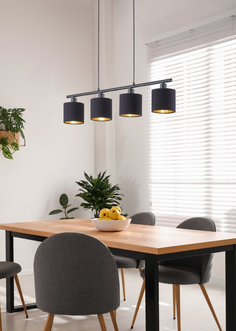 Een moderne eetkamer met een houten tafel, vier grijze stoelen en een zwarte hanglamp erboven. Er staat een schaal met gele appels op tafel. Er staat een potplant in de hoek en er stroomt licht door witte jaloezieën.