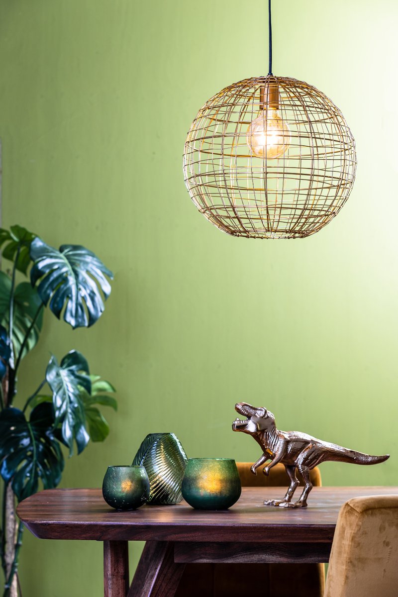 Een kamer met een moderne, bolvormige hanglamp van draad boven een houten tafel. Op de tafel staan twee groene glazen vazen en een klein dinosaurusbeeldje. Een grote bladplant staat naast de tafel tegen een groene muur.