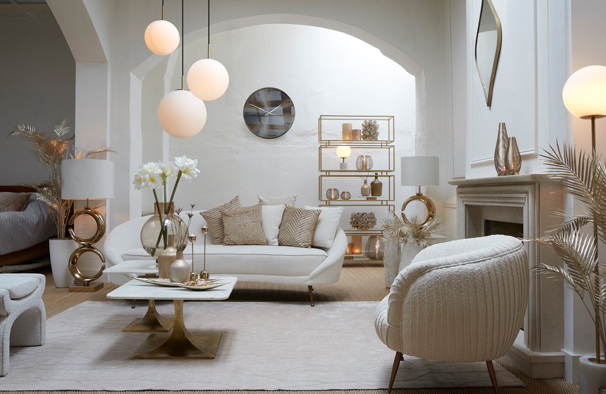 Een stijlvolle woonkamer met een moderne inrichting met witte banken, een ronde fauteuil en een marmeren salontafel. Gouden accenten zijn onder andere een plankenkast, decoratieve items en verlichting. Een neutraal tapijt bedekt de vloer en planten zorgen voor een vleugje groen.