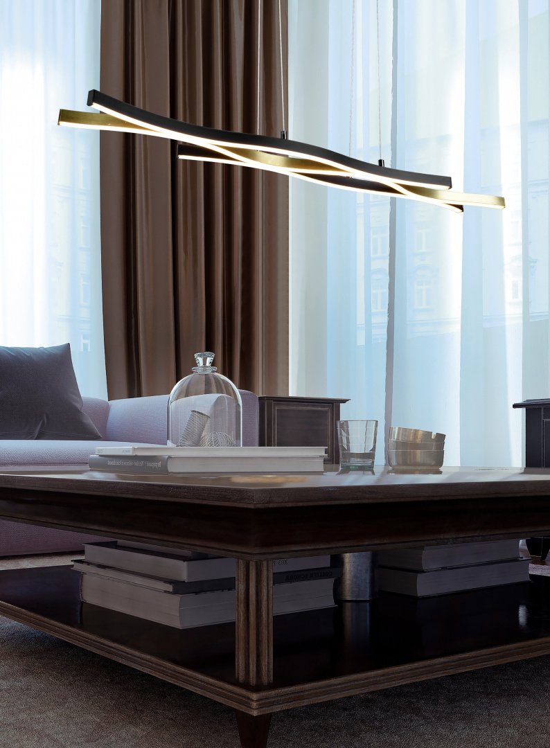 Een moderne woonkamer met een strakke zwarte kroonluchter boven een houten salontafel. De tafel bevat een glazen koepel, twee metalen containers en verschillende boeken. Een grijze bank staat op de achtergrond, met doorschijnende en bruine gordijnen die grote ramen bedekken.