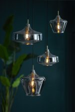 Vier moderne hanglampen met heldere glazen kappen hangen op verschillende hoogtes, elk met een spiraalvormige gloeilamp. De achtergrond is diepblauwgroen, geaccentueerd door het zachte silhouet van bladplanten aan de linkerkant.