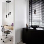 Moderne kantoorruimte met een strakke grijze stoel aan een houten bureau onder hangende lampen. Een gesloten laptop en een decoratief figuur staan op het bureau. In de buurt staan een zwarte kast en kom bij een raam met zwarte jaloezieën. Neutrale tinten creëren een minimalistische sfeer.