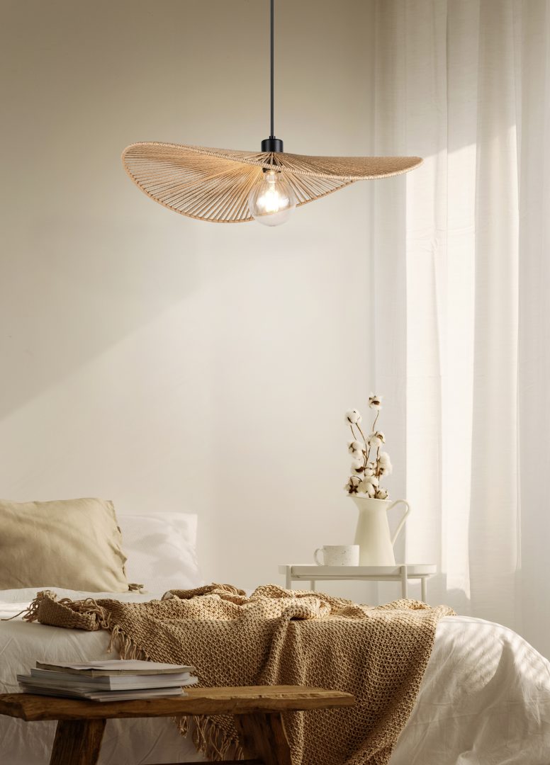 Een gezellige slaapkamer met een beige kleurenschema heeft een bed met een gebreide deken en kussens. Een unieke, sculpturale hanglamp hangt aan het plafond. Een kleine houten bank staat aan het voeteneind van het bed en zacht natuurlijk licht filtert door transparante gordijnen.