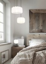Een gezellige slaapkamer met een houten bed, gebroken wit beddengoed en een grof gebreide deken. Twee hanglampen hangen boven een houten nachtkastje met een klein lampje. Er hangt een grote spiegel met houten lijst aan de muur en er is een raam met witte gordijnen.