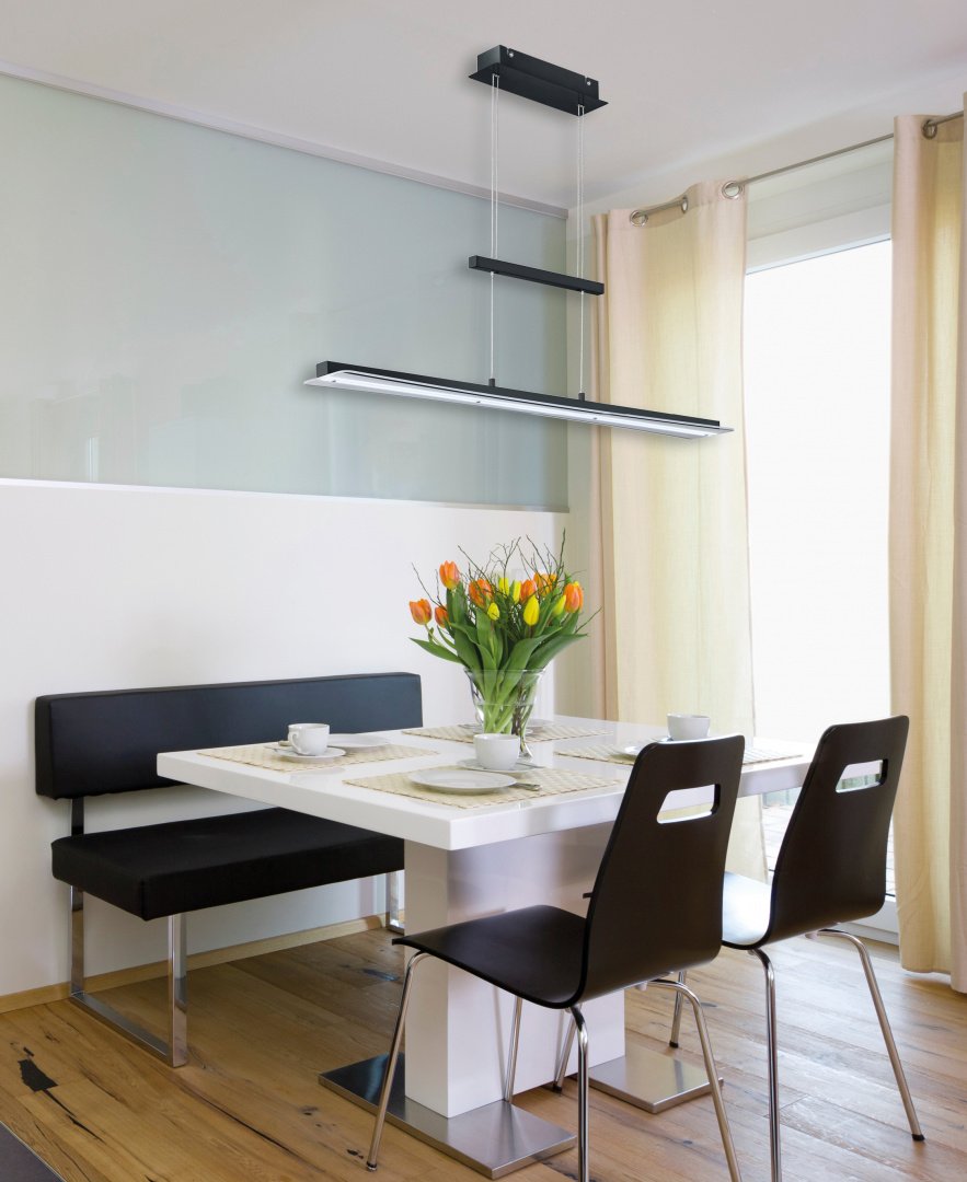Een moderne eethoek met een witte tafel en twee donkere stoelen, een zwarte bank met kussens en een vaas met oranje tulpen. Een strakke zwarte hanglamp hangt erboven. Grote ramen met beige gordijnen laten natuurlijk licht de ruimte vullen.