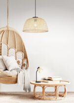 Een gezellige kamer met een hangende rieten stoel met kussens en een kleine rotan tafel. Een open boek ligt op de tafel met een kleine lamp. Een grote, geweven hanglamp hangt aan het plafond en de kamer heeft een neutraal kleurenpalet.