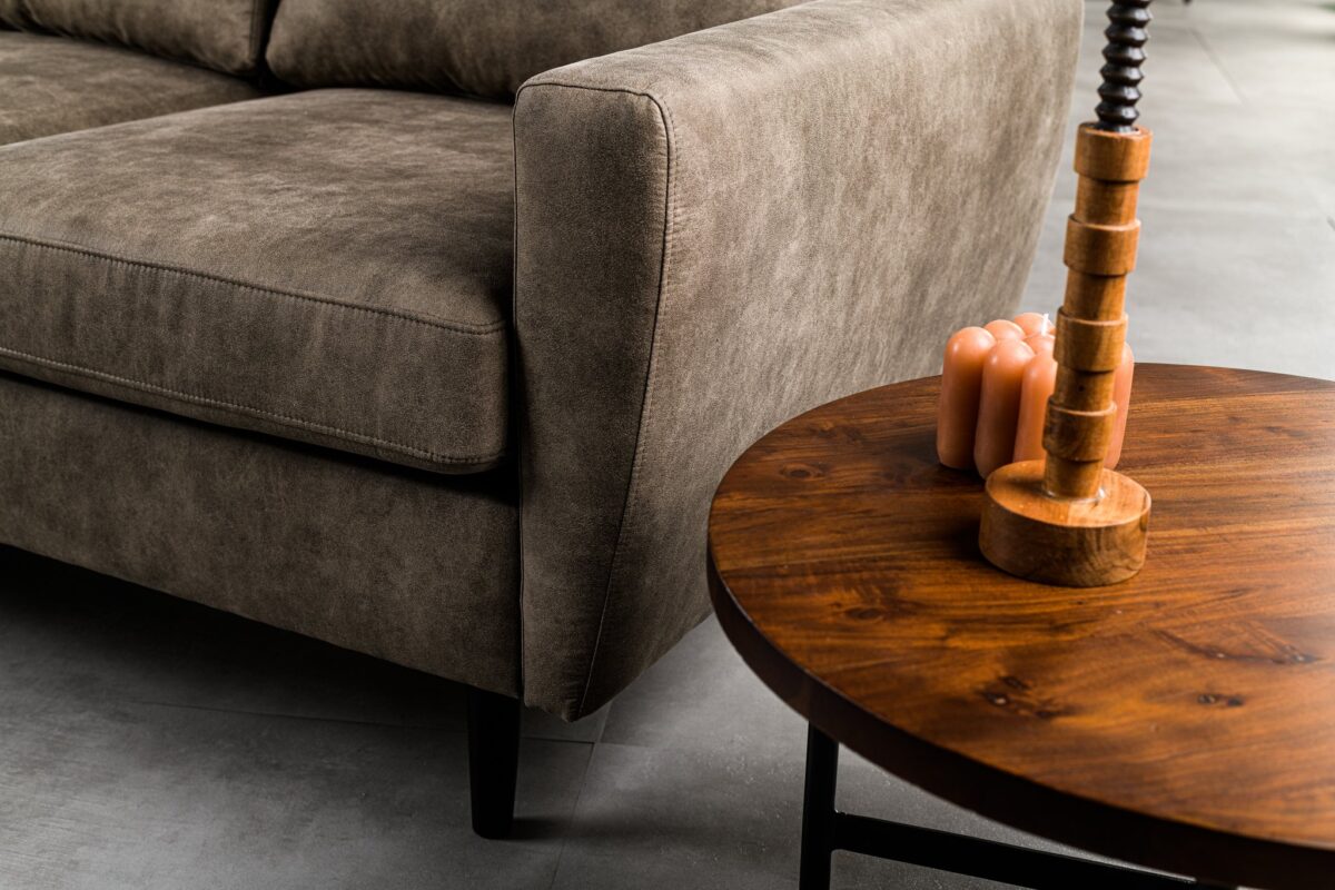 Een close-up van een moderne woonkamer met een bruine stoffen bank en een ronde houten salontafel. Op de tafel staat een houten lamp met een spiraalvormige voet en een kleine decoratieve kaars, op een gladde, grijze vloer.