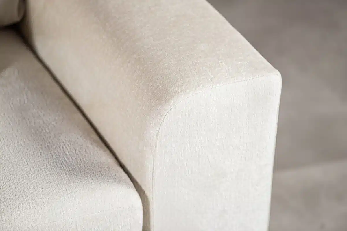 Close-up van een beige armleuning van een bank, met een getextureerde stof. De rand is glad met zachte rondingen en de achtergrond is zacht vervaagd, waardoor de details van de bekleding worden benadrukt.