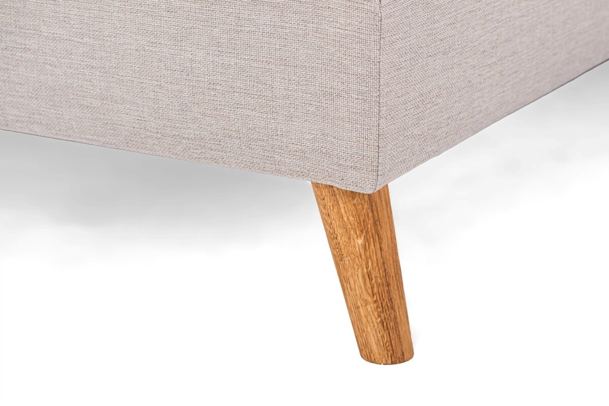 Close-up van een beige stoffen hoekbank met een enkele houten poot op een witte achtergrond. De stof heeft een subtiel structuurpatroon en de houten poot is taps toelopend en lichtbruin.