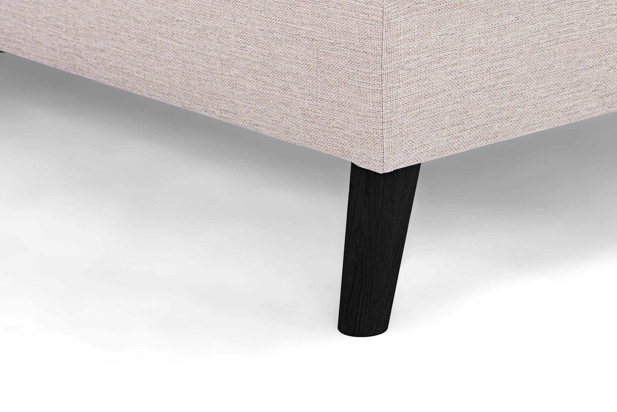 Close-up van een beige gestoffeerde hoekbank met structuurstof en een strakke zwarte houten poot. De achtergrond is een eenvoudig wit oppervlak.