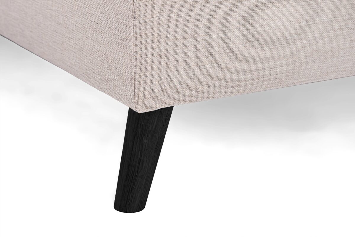 Close-up van een beige gestoffeerde hoekbank met een getextureerde stof. De bank heeft strakke zwarte houten poten en staat tegen een effen witte achtergrond.