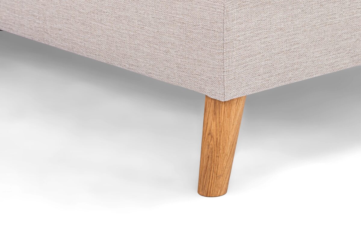 Close-up van een beige stoffen hoekbank met een enkele houten poot op een lichte achtergrond. De stof heeft een subtiel gestructureerd patroon en de poot is een gladde, natuurlijke houtafwerking, die contrasteert met de bekleding.