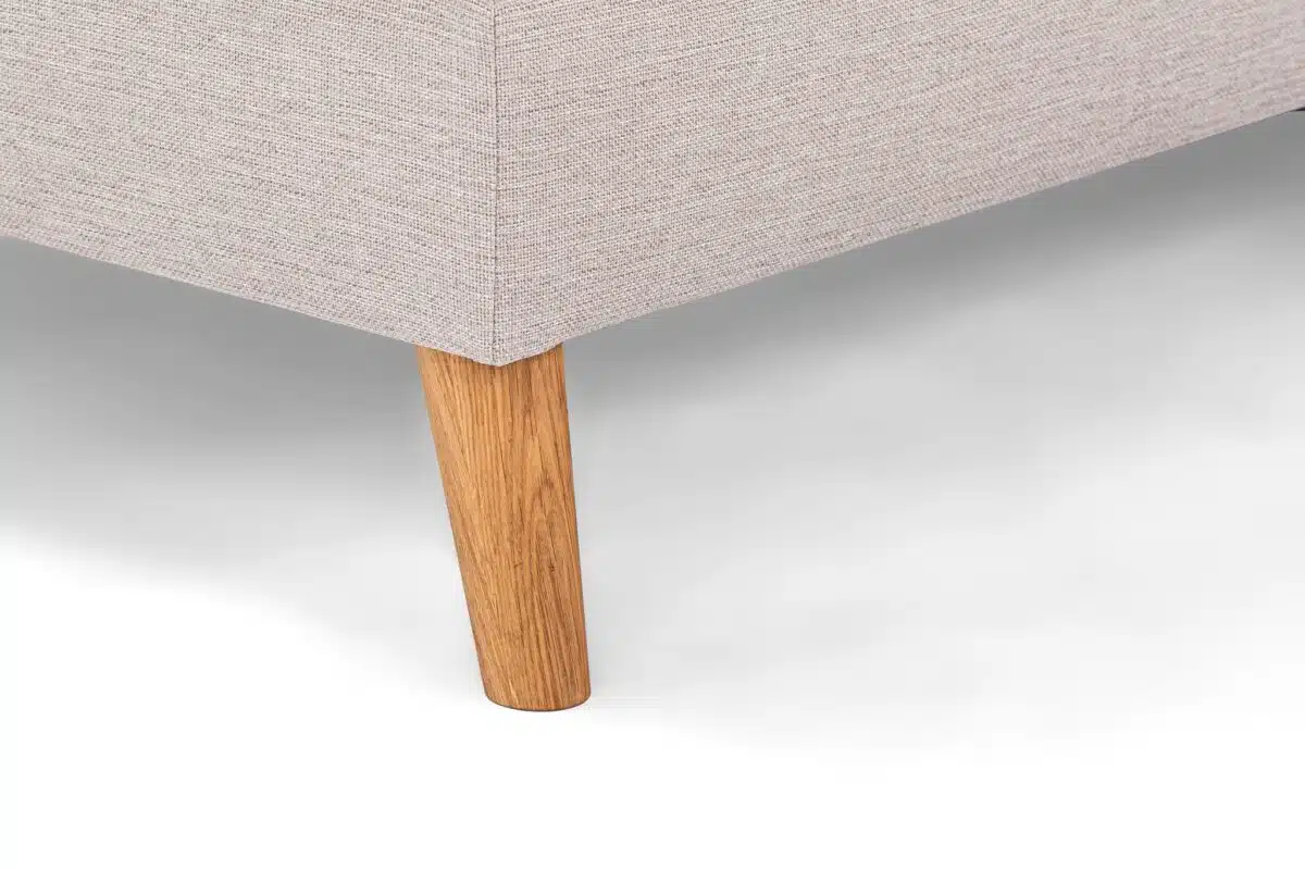 Close-up van een beige hoekbank met structuurstof en een enkele houten poot. De poot is taps toelopend en lichtbruin, en loopt door vanaf de onderkant van de bank, die op een wit oppervlak staat.