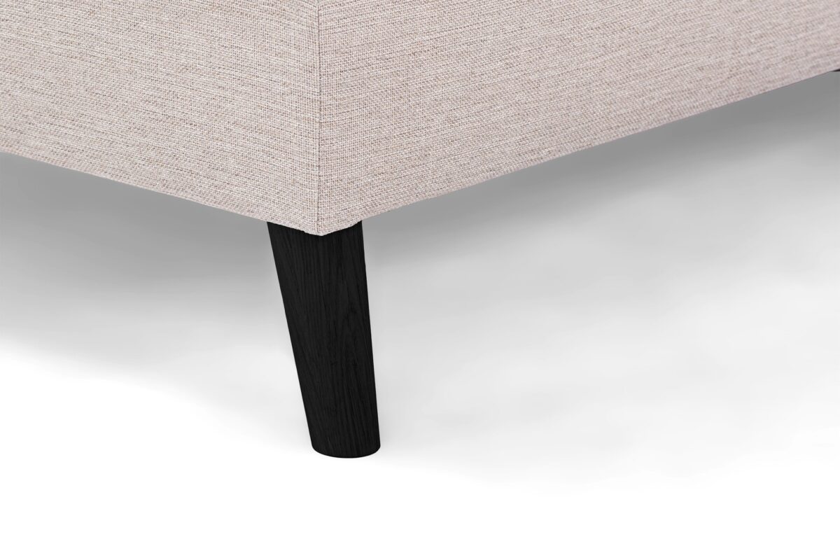Close-up van een beige stoffen hoekbank met de structuurbekleding en één enkele zwarte houten poot op een witte achtergrond.