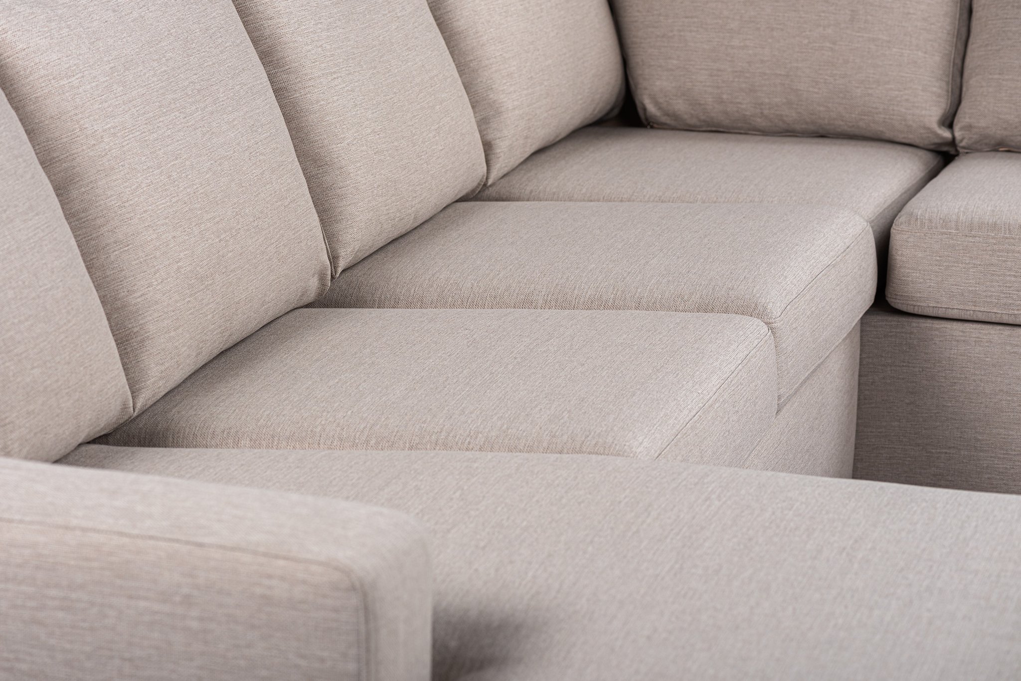 Close-up van een beige sectionele bank met het hoekgedeelte en zachte zit- en rugkussens. De stof van de bank ziet er glad en zacht uit, wat zorgt voor een comfortabele en uitnodigende look.