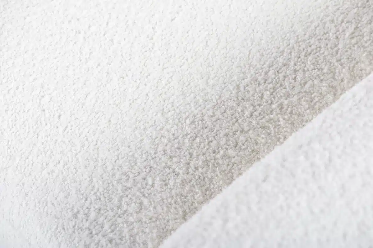 Close-up van een zachte, gestructureerde witte stof met een pluizige, wolachtige uitstraling. De stof is in zachte vouwen gerangschikt, waardoor er subtiele schaduwen over het oppervlak werpen en de pluche textuur wordt benadrukt.