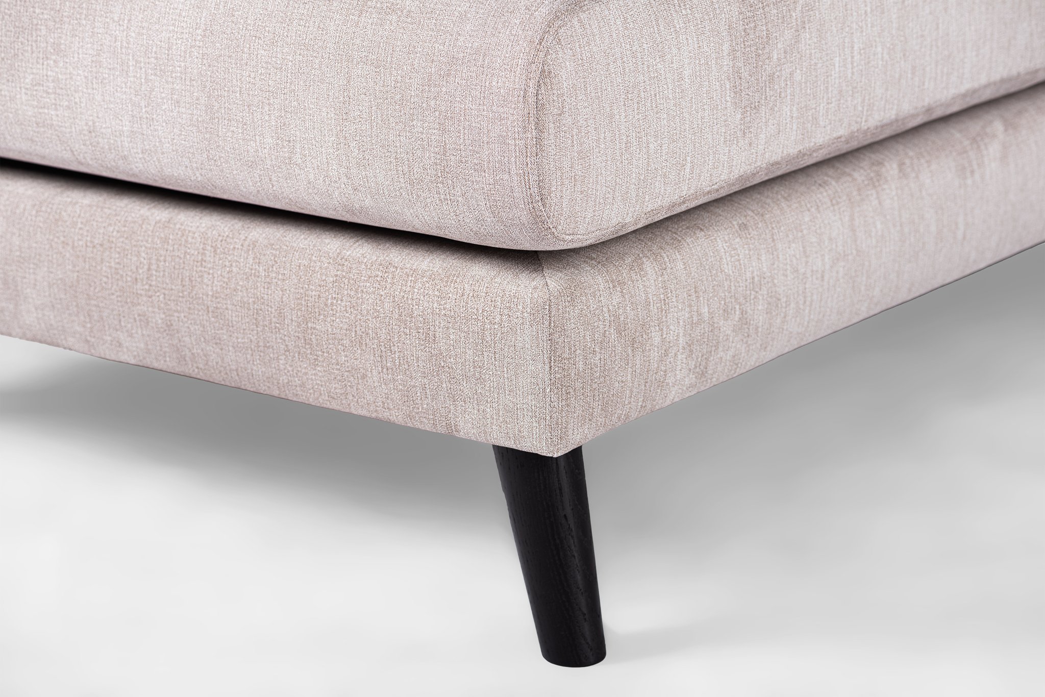 Close-up van een lichtgrijze gestoffeerde hoekbank met gladde stoffen textuur. De bank wordt ondersteund door een strakke, taps toelopende zwarte houten poot, die contrasteert met de lichte bekleding. De afbeelding legt de elegantie en eenvoud van modern meubeldesign vast.
