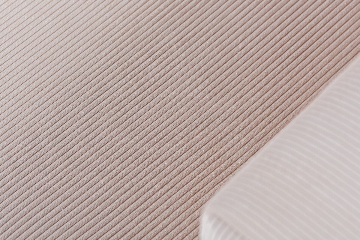 Close-up van een gestructureerd stoffen oppervlak met diagonale lijnen in beige en een witte hoek met fijne strepen. De stof creëert een subtiel patroon, wat suggereert dat het een zacht en comfortabel textielontwerp is.
