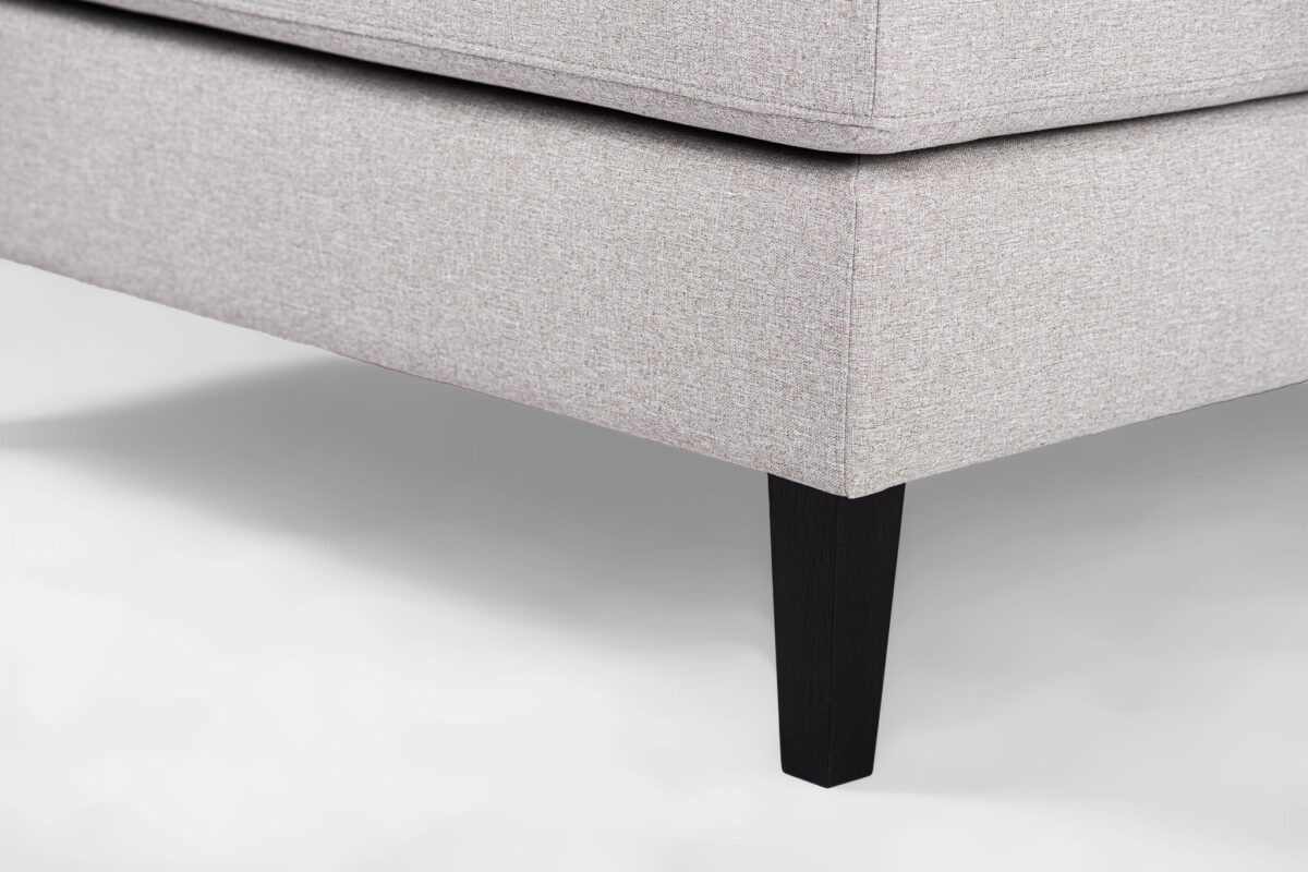 Close-up van een beige stoffen hoekbank met strakke lijnen en modern design. Het heeft een gladde afwerking en rust op slanke, donkere houten poten, die subtiel contrasteren met de bekleding. De achtergrond is een zachte, lichte kleur.