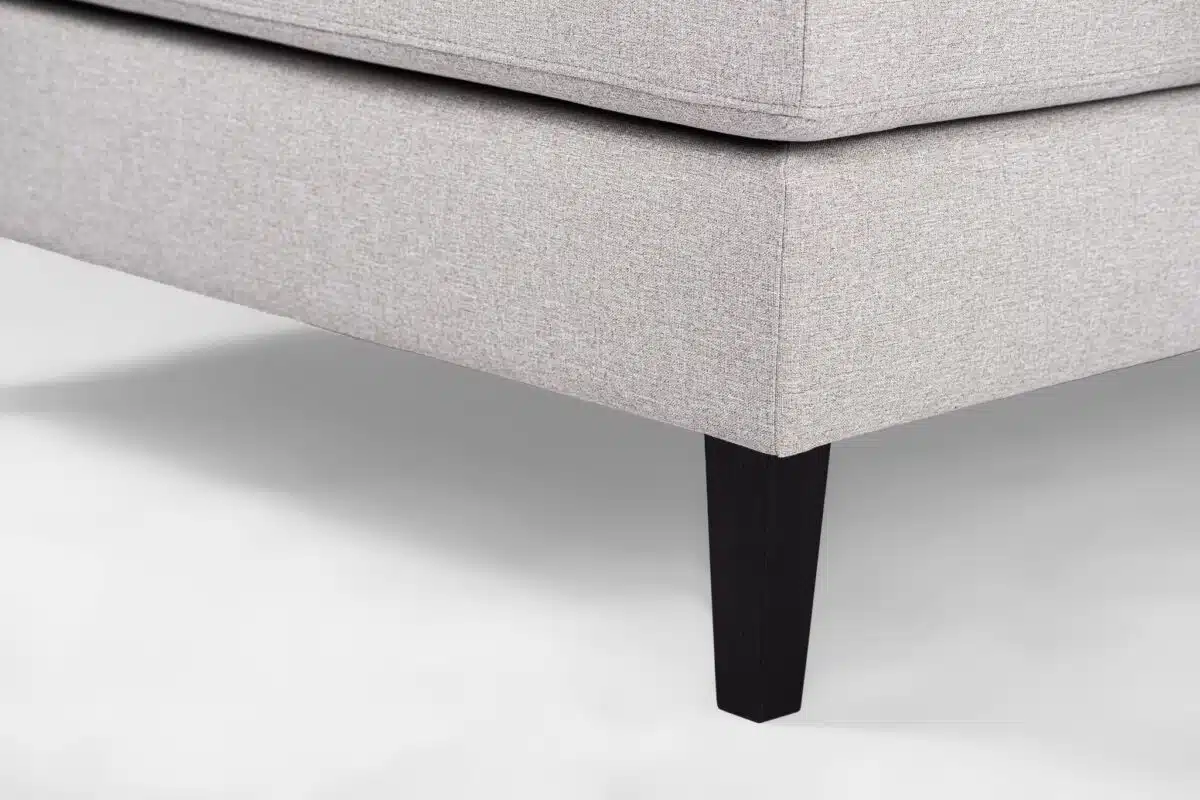 Close-up van een beige stoffen hoekbank met strakke lijnen en modern design. Het heeft een gladde afwerking en rust op slanke, donkere houten poten, die subtiel contrasteren met de bekleding. De achtergrond is een zachte, lichte kleur.