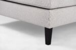 Close-up van een beige stoffen hoekbank met strakke lijnen en modern design. Het heeft een gladde afwerking en rust op slanke, donkere houten poten, die subtiel contrasteren met de bekleding. De achtergrond is een zachte, lichte kleur.