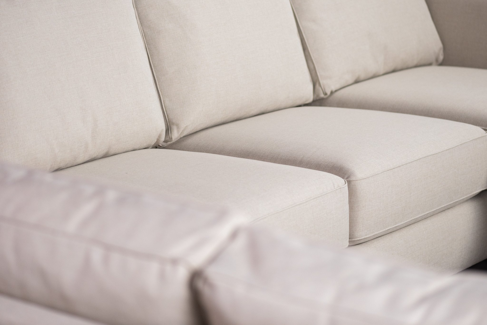 Close-up van een beige bank met pluche kussens. De stof ziet er glad en zacht uit en de zitting ziet er ruim en comfortabel uit. De strakke lijnen en neutrale kleur van de bank geven hem een moderne en minimalistische uitstraling.